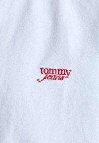 Hvit stoff med en rød brodert logo som leser "tommy jeans." Teksturen er glatt med en svak glans.