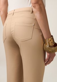 Beige broek met een gladde textuur heeft twee achterzakken en subtiele stiksels. Een gouden armband siert de pols van het model.