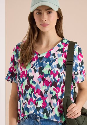 Mujer con camisa de estampado abstracto multicolor, gorra verde claro y lleva una correa de bolso de hombro acolchada verde oscuro.