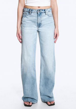 Personne portant un jean taille haute, bleu clair, coupe large, un top court blanc et des sandales noires ouvertes aux orteils, debout sur un fond blanc.