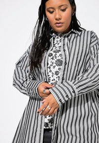 Camicia a maniche lunghe a righe bianco e nero con chiusura a bottoni, caratterizzata da una stampa floreale sottostante e un design di polsini a contrasto.