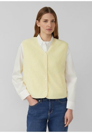 Kobieta o długich brązowych włosach, ubrana w bladożółtą kamizelkę z polaru zapinaną na guziki, na białej koszuli i niebieskich dżinsach, stoi z jedną ręką w kieszeni.