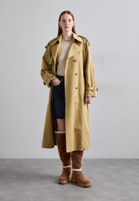 Beige trench coat with double-breasted buttons, wide lapels, and shoulder epaulettes; paired with a grey sweater and denim skirt. Brown shearling-lined boots.