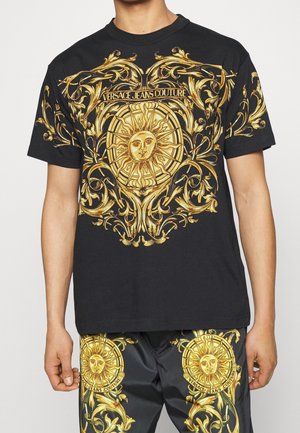 Mies pukeutuneena mustaan Versace Jeans Couturen asuun, jossa on kultaiset, koristeelliset auringon ja käärmeiden kuviot paidassa ja housuissa.