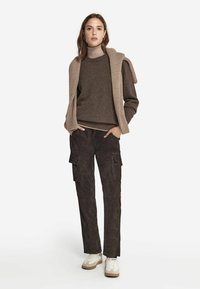 Pull en laine brun texturé sur un col roulé beige, associé à un pantalon cargo noir avec des poches, et des baskets blanches avec des accents beiges.