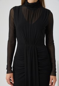 Μαύρο φόρεμα με μακριές διαφανείς μανίκες, συγκεντρωμένο μπούστο, ψηλό λαιμό και ρέουσα λεπτομέρεια στην μέση. Απαλή υφή υφάσματος.
