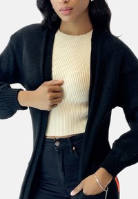 Czarny dziergany kardigan z otwartym frontem, długimi rękawami i ściągaczami przy mankietach. Nosi się go na kremowy, prążkowany top, w połączeniu z czarnymi jeansami.