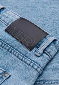 Tela de denim azul claro con una superficie texturizada y una etiqueta de cuero negra con texto en relieve visible en el bolsillo trasero.