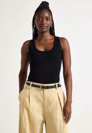 Punottu hius nainen, joka käyttää mustaa hihattomaa yläosaa, beigejä laskostettuja housuja ja mustaa vyötä, poseeraa yksinkertaisen valkoisen taustan edessä.