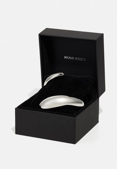 Dva srebrna metalna predmeta s elegantnim, zakrivljenim dizajnom unutar kutije obložene crnim baršunom, označeni s "HUGO KREIT." Kutija ima mat završnu obradu.