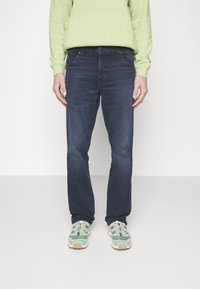 Vaqueros de denim azul oscuro con un corte recto, ligeras decoloraciones y diseño clásico de cinco bolsillos, combinados con zapatillas verdes y beige.