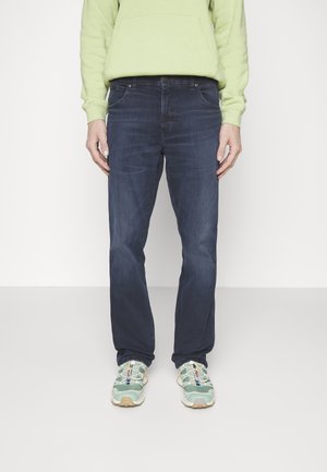 Dunkelblaue Jeans aus Denim mit geradem Bein, dezentem Verblassen und klassischem Fünf-Taschen-Design, kombiniert mit grünen und beigen Sneakers.