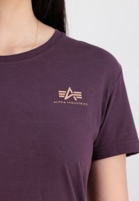 Lilla bomulds t-shirt med korte ærmer, rund halsudskæring og et lille metallisk guld logo med en stjerne og tekst på brystet.