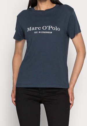 Kobieta ubrana w granatowy t-shirt z "Marc O'Polo EST. IN STOCKHOLM" białym tekstem, w połączeniu z czarnymi spodniami, stojąca na białym tle.