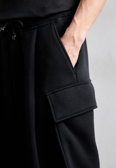 Black joggers made of soft fabric, featuring a side pocket and an elastic waistband with drawstring. Smooth texture and minimalist design.