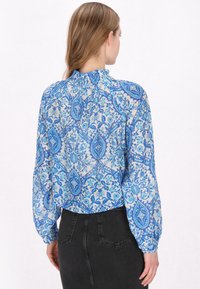 Blaue gemusterte Bluse mit langen Ärmeln, gerafftem Ausschnitt und einer Mischung aus floralen und geometrischen Designs auf einem transparenten Stoff. Kombiniert mit schwarzen Jeans.