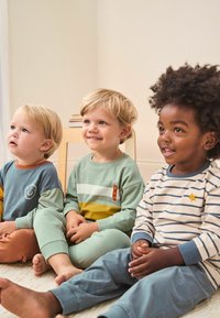 Puha pamutból készült gyerek otthoni ruházat: az egyik zöld és kék árnyalatokat visel mosolygós mintával, a másik csíkos bézsben csillag díszítéssel.