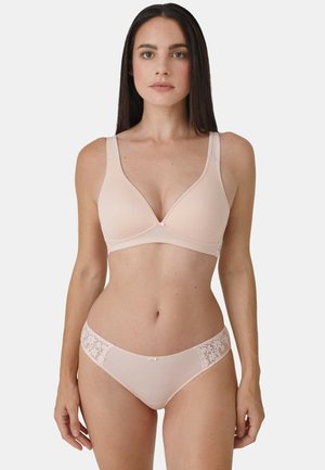 Frau mit langen dunklen Haaren, die einen hellbeigen BH und passende Unterwäsche mit Spitzenakzenten trägt, steht vor einem schlichten hellen Hintergrund.