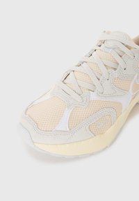 Jasnobeżowe sneakersy z siateczki i zamszu, o zaokrąglonym kształcie, teksturowanej powierzchni i białych akcentach. Sznurówki są zawiązane i widoczne.