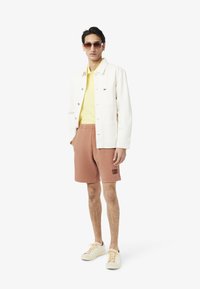 Chaqueta blanca, polo amarillo, pantalones cortos rosas con logo y zapatillas crema. Las gafas de sol completan el look. Casual, ajuste moderno.