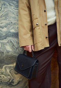 Čierna kožená kabelka s textúrovaným povrchom, obdĺžnikového tvaru a vrchným uchom, ktorú drží osoba v béžovej bunde a hnedých nohaviciach.