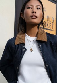 Τζιν μπουφάν με γιακά από κορντόρι, λευκό πουκάμισο από κάτω, στρωμένα χρυσά κολιέ και απλά σκουλαρίκια καρφωτά, τοποθετημένα σε ανοιχτό φόντο.