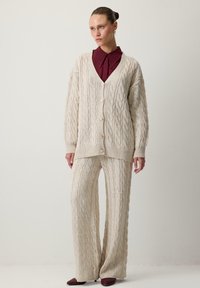 Beige kabelgebreide cardigan met een V-hals en knopen, gecombineerd met bijpassende wijde broek. Zachte textuur, contrasterend bordeauxrood onderhemd.