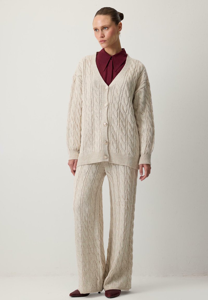 Beige kabelgebreide cardigan met een V-hals en knopen, gecombineerd met bijpassende wijde broek. Zachte textuur, contrasterend bordeauxrood onderhemd.