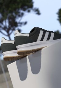 Zapatillas Adidas en blanco y negro con tres rayas, exhibidas sobre una superficie blanca al aire libre con árboles desenfocados al fondo.