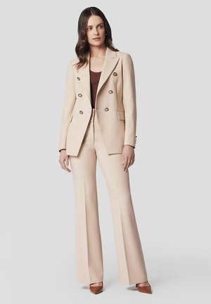 Beżowa marynarka szyta na miarę z podwójnymi złotymi guzikami, zestawiona z pasującymi spodniami z wysokim stanem. Brązowa bluzka i czó�łenka na kwadratowym nosku dopełniają całość.