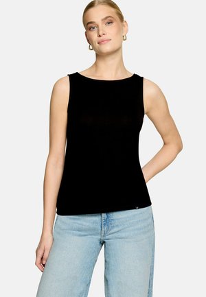 Femme portant un haut noir sans manches et un jean bleu clair, debout avec une main sur la hanche sur un fond uni.