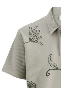 Grijze geribbelde polo met zwarte geborduurde bloemen- en handontwerpen. Voorzien van een klassieke kraag en korte mouwen.