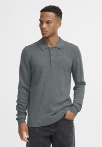 Grijze gebreide polo trui met lange mouwen en een kraag, drie-knopen sluiting en geribbelde manchetten. Draagt met donkere jeans.