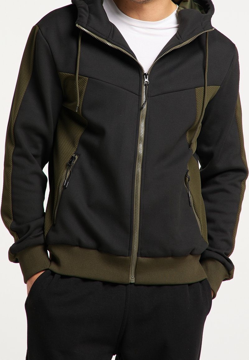 Homme portant un sweat à capuche zippé noir et vert olive avec des poches zippées à l'avant et un T-shirt blanc en dessous.