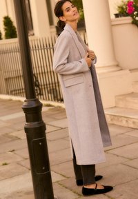 Un manteau en laine grise clair mélangée avec des revers en notch et deux poches, associé à un jean foncé et des chaussures plates noires, se tenant près d'un lampadaire.
