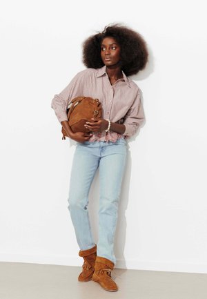 Femme avec un afro volumineux portant une chemise rayée, un jean bleu clair, des bottes en daim marron, tenant un sac à main en daim marron contre un mur blanc.