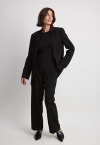 Schwarzer, taillierter Blazer mit scharfen Revers, kombiniert mit einem figurbetonten schwarzen Oberteil und weiten schwarzen Hosen, akzentuiert durch spitze schwarze Pumps.