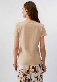Camiseta de algodón beige con mangas cortas, cuello redondo y un pequeño logo en la parte trasera. Combinada con pantalones estampados y de corte holgado.