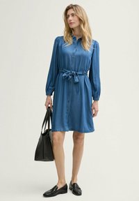 Denim shirt dress in medium blue with long puffed sleeves, a button-down front, a belted waist, and a flared hem. Black tote bag and loafers.