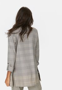 Vrouw met lang donker haar, met de rug naar de kijker, draagt een grijs-wit geruit colbert met opgerolde mouwen en bijpassende geruite broek.