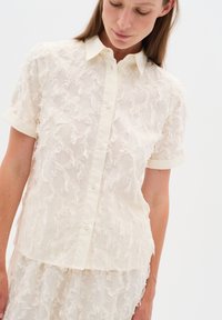 Cream-colored blouse with a textured fringe design, short sleeves, and a standard collar, featuring a button-down front.
