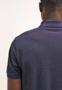 Homme portant un polo bleu foncé à manches courtes, vu de dos sur un fond clair uni.