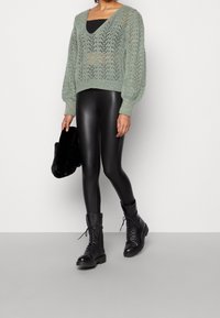 Pull vert clair en maille avec col en V et manches bouffantes, associé à des leggings noirs très brillants et des bottines de combat noires à lacets.