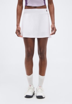 Vrouw draagt een witte sportrok, witte sokken en witte sneakers, staand tegen een effen achtergrond.