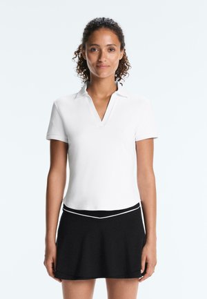 Kvinde stående med fronten mod kameraet iført en hvid kortærmet skjorte med krave og et sort skørt med hvid kant.