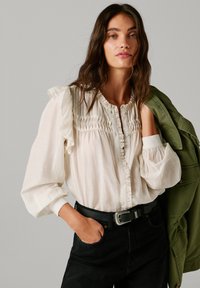 Balta, raukšlėta bluzka su mygtukų priekyje ir tekstūriniu suraukimu; dėvima su juodu diržu ir tamsiomis džinsomis, derinama su alyvmedžio žalia striuke.