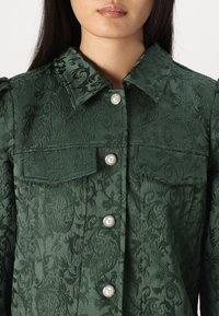Mörkgrön jacka med blomstrande texturmönster, med krage, två bröstfickor och dekorativa pärltryckknappar.