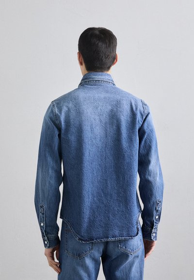 Camicia in denim di blu medio a scuro, con design a bottone, maniche lunghe e orlo curvo. Tessuto testurizzato con cuciture visibili.