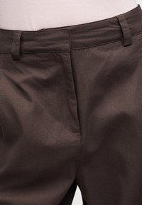 Κοντινή φωτογραφία στη μέση σκούρων καφέ παντελονιών με θηλιές για ζώνη και κρυφό μπροστινό κούμπωμα πάνω σε ανοιχτόχρωμο πουκάμισο.