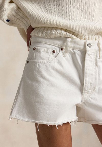 Λευκά τζιν σορτς με ξεφτισμένα τελειώματα, φορεμένα με εκρού πλεκτό πουλόβερ, το χέρι ακουμπισμένο στην τσέπη, σε φωτεινό φόντο.